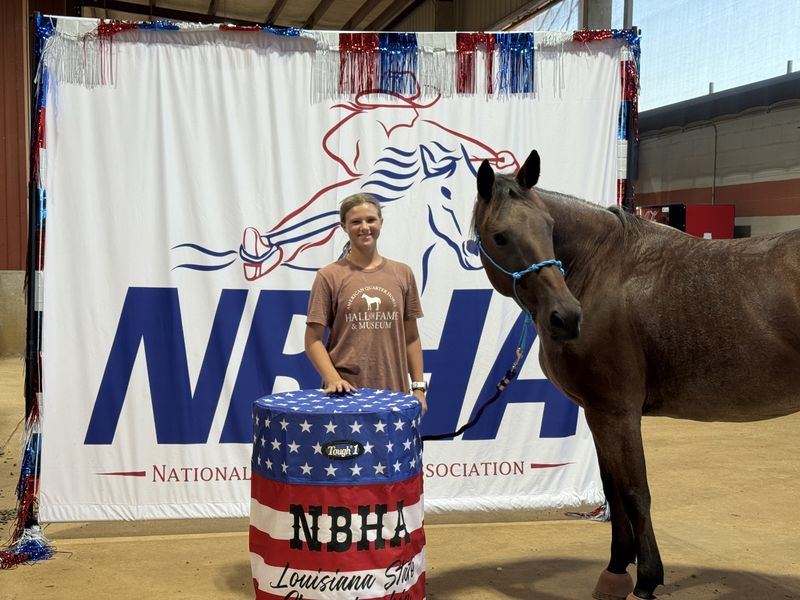 Adeline Frost smiling beside Pepper, her red roan horse, in front of the NBHA Louisiana State backdrop and barrel